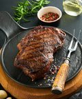 Grilled steak on a black plate with a wooden cutting board, fork, and garnishes.