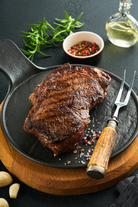 Grilled steak on a black plate with a wooden cutting board, fork, and garnishes.