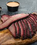 Sliced brisket on a wooden cutting board with side dishes of sauces in the background.