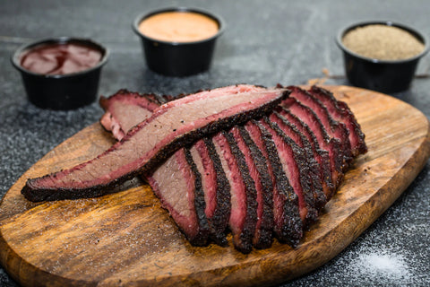 Sliced brisket on a wooden cutting board with side dishes of sauces in the background.