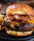 Hamburger with bacon and cheese on a bun with waffle fries and ketchup in the background.