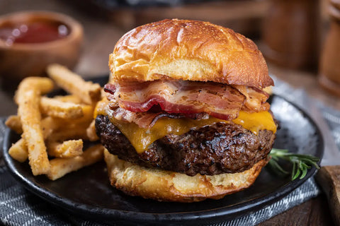 Hamburger with bacon and cheese on a bun with waffle fries and ketchup in the background.