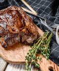 Grilled steak on a wooden cutting board with herbs and a bowl of peppercorns on a checkered cloth.