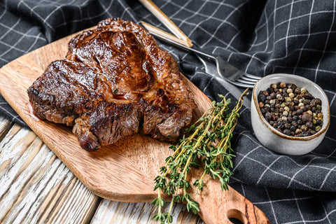 Grilled steak on a wooden cutting board with herbs and a bowl of peppercorns on a checkered cloth.