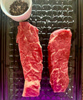Two raw steaks on a metal tray with a small bowl of spices.