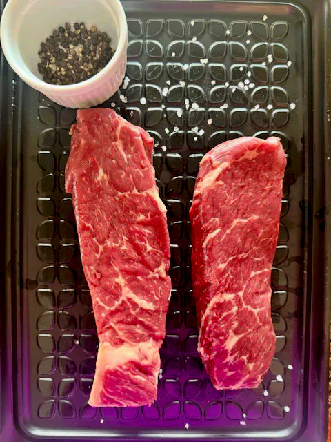 Two raw steaks on a metal tray with a small bowl of spices.