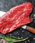 Raw beef steak on a dark surface with a knife, rosemary, and salt.