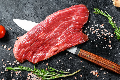 Raw beef steak on a dark surface with a knife, rosemary, and salt.