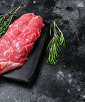 Raw steak on a black slate board with herbs and red pepper flakes on a dark surface