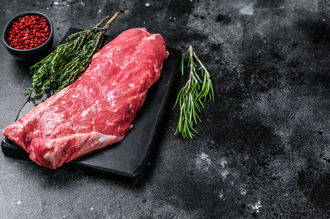 Raw steak on a black slate board with herbs and red pepper flakes on a dark surface