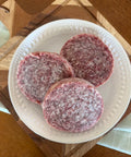 Three raw hamburger patties on a white plate with a wooden surface underneath.