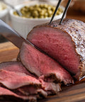 Sliced roast beef on a wooden cutting board with a knife and fork.
