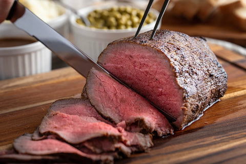 Sliced roast beef on a wooden cutting board with a knife and fork.