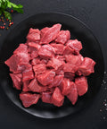 Raw beef chunks on a black plate with herbs and spices on a dark surface