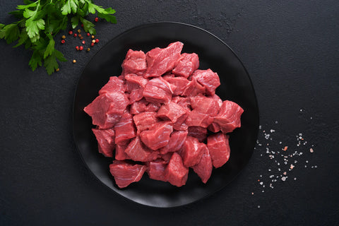 Raw beef chunks on a black plate with herbs and spices on a dark surface