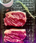 Two raw steaks on a metal surface with rosemary and a small container of spices.