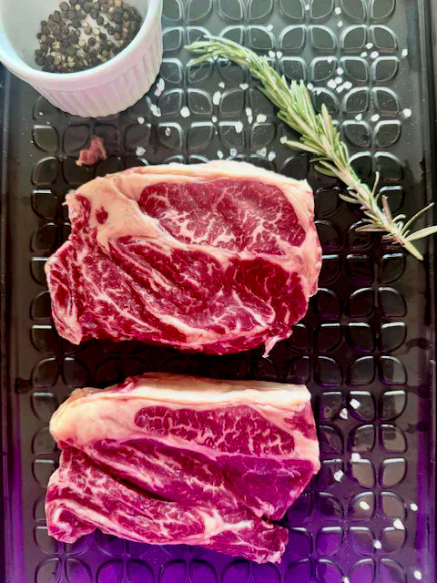 Two raw steaks on a metal surface with rosemary and a small container of spices.