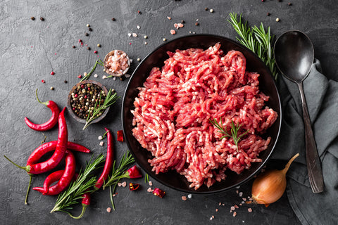 Ground meat on a black plate with red peppers, rosemary, and a spoon on a dark surface.
