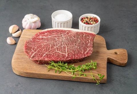 Raw steak on a wooden cutting board with garlic, salt, and peppercorns on a gray background