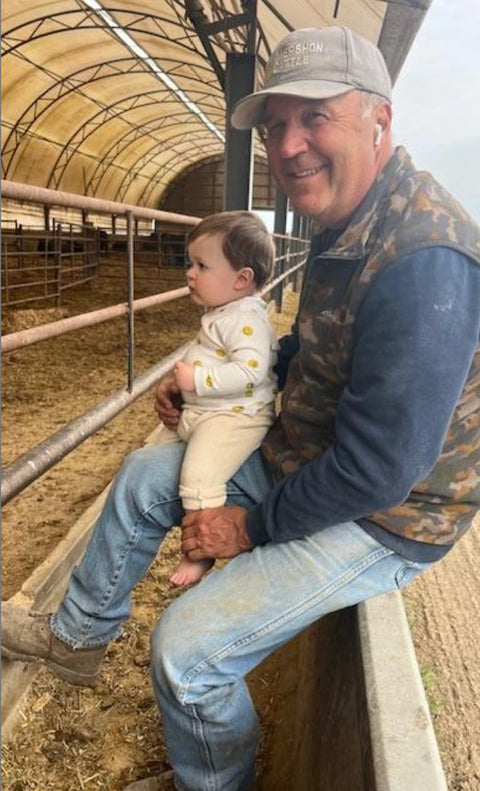 A smiling man wearing a gray cap, blue jeans, and a camouflage vest sits on a concrete barrier in a barn, holding a baby dressed in a cream outfit with yellow polka dots.