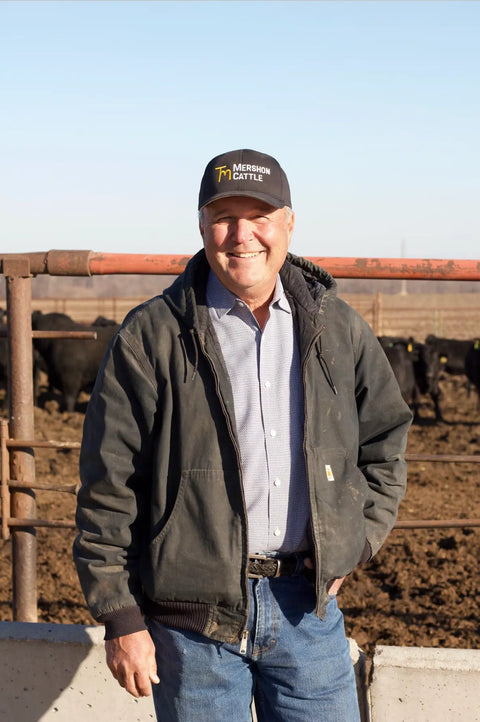 Man in black jacket and blue jeans with Mershon Cattle hat standing on a farm with cows 