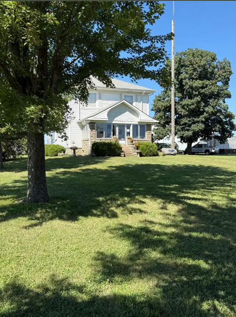 A white two-story house with a front porch and stone steps, set on a grassy lawn with large green trees under a blue sky.