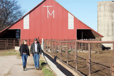 Two people walking in front of a red barn with a silo in the background.