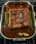 A large roasted beef brisket sits in a stainless steel All-Crocs roasting pan on a gas stovetop, surrounded by browned fat, rich brown sauce, rosemary sprigs, and bay leaves.