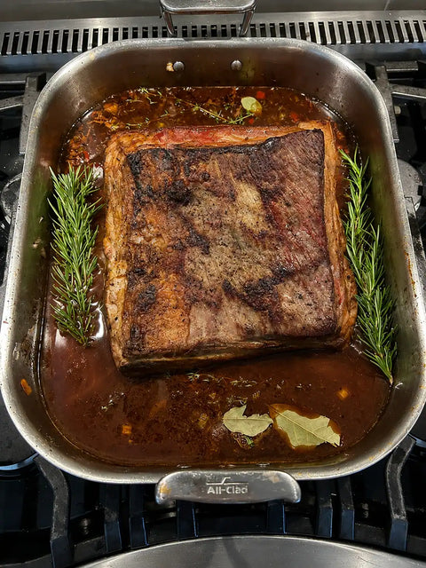 A large roasted beef brisket sits in a stainless steel All-Crocs roasting pan on a gas stovetop, surrounded by browned fat, rich brown sauce, rosemary sprigs, and bay leaves.