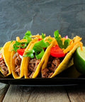 Three tacos with ground beef, lettuce, and tomato on a black plate with a lime wedge on a wooden surface.