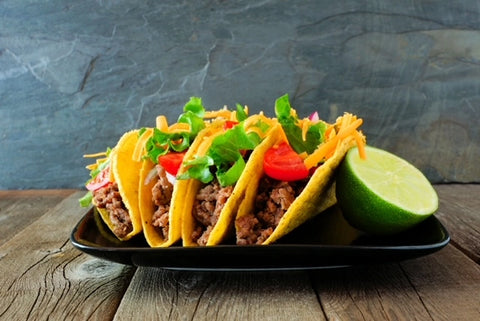 Three tacos with ground beef, lettuce, and tomato on a black plate with a lime wedge on a wooden surface.