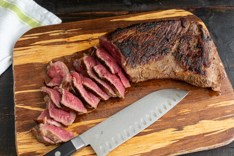 Beef tri-tip cooked and sliced on a wooden cutting board next to a knife