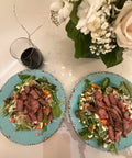 Two plates of salad with beef and vegetables on a marble table with flowers and a glass of red wine.