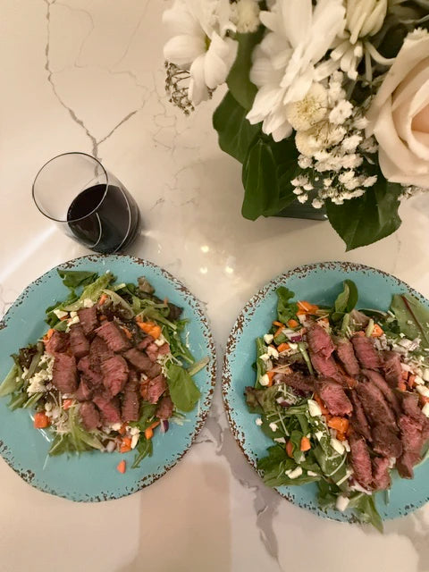 Two plates of salad with beef and vegetables on a marble table with flowers and a glass of red wine.